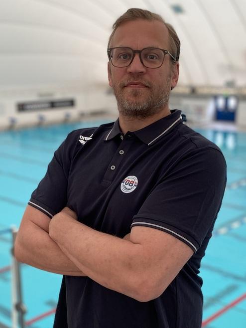 Mann mit Brille und Polo-Shirt steht an einem Indoor-Schwimmbecken mit blauer Wasseroberfläche im Hintergrund.